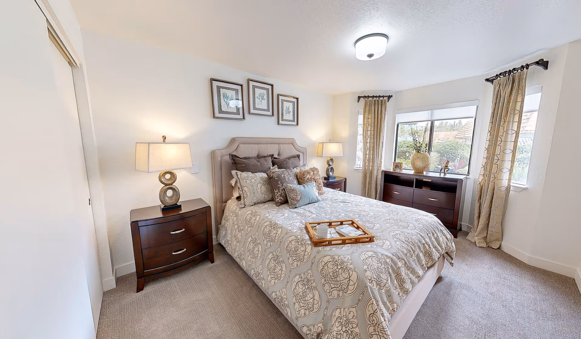 A well-lit bedroom with a large bed covered in a patterned beige and white bedspread, multiple decorative pillows, and a wooden tray with a cup and book. There are two dark wooden nightstands with modern lamps on either side of the bed. Three framed botanical prints hang above the bed. The room has beige carpet, two windows with beige patterned curtains, and a dark wooden dresser with decorative items on top.