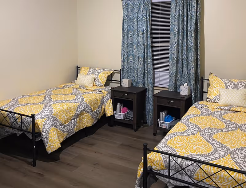 A bedroom with two single beds featuring yellow and gray patterned bedding. Between the beds are two dark wooden nightstands, each holding a tissue box and a small basket with personal care items. A window with blue patterned curtains is centered on the wall behind the nightstands.