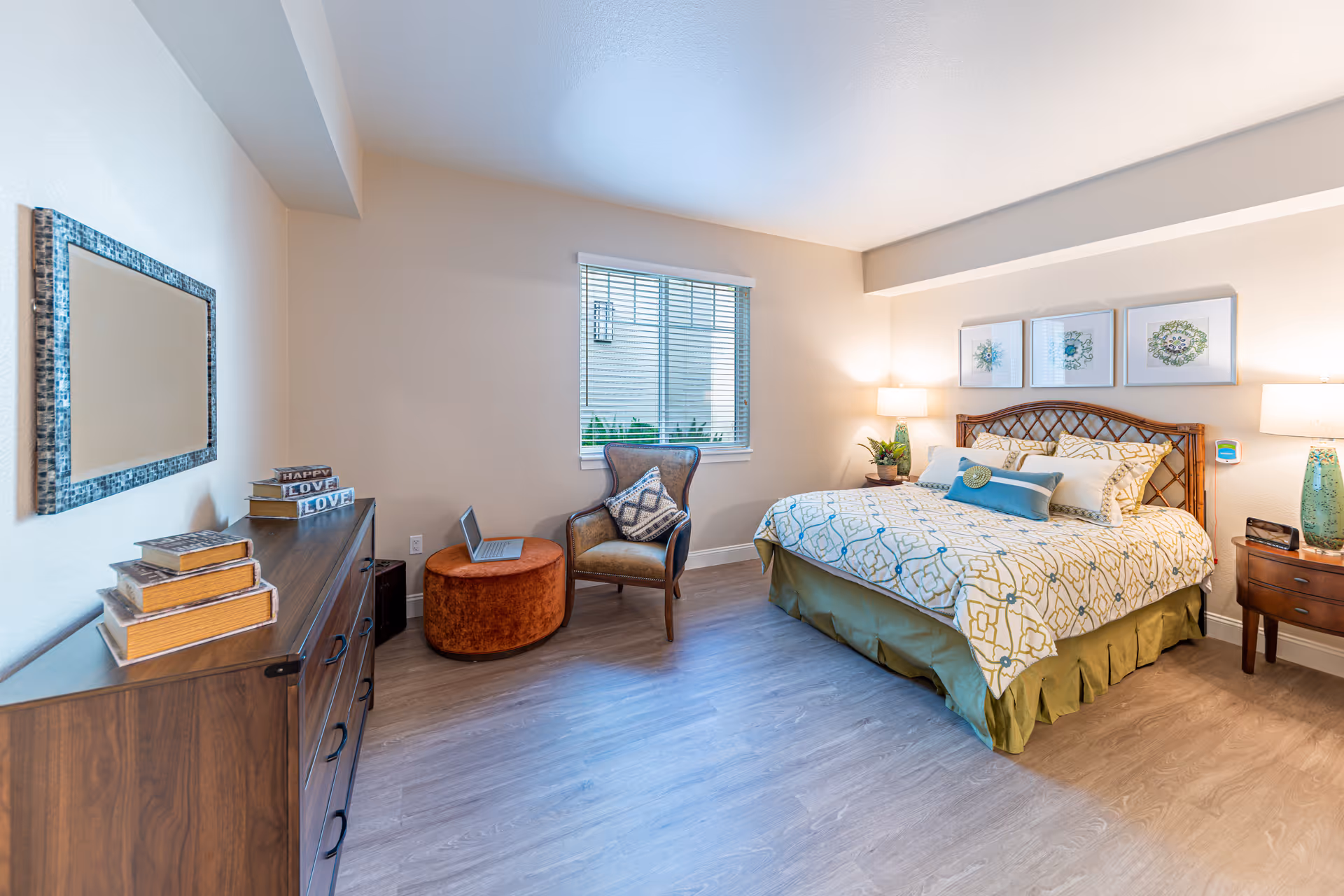 A bright and cozy bedroom featuring a large bed with patterned bedding and multiple pillows, two bedside tables with lamps, a wooden dresser with decorative books, a cushioned chair with a pillow, and a round ottoman with a laptop on it. A window with blinds lets in natural light, and three framed floral artworks hang above the bed.