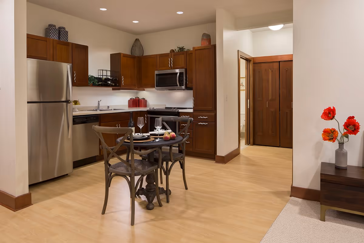 Open kitchen and dining area with stainless steel appliances, wooden cabinets, a small round table with two chairs, and a side table holding red flowers.