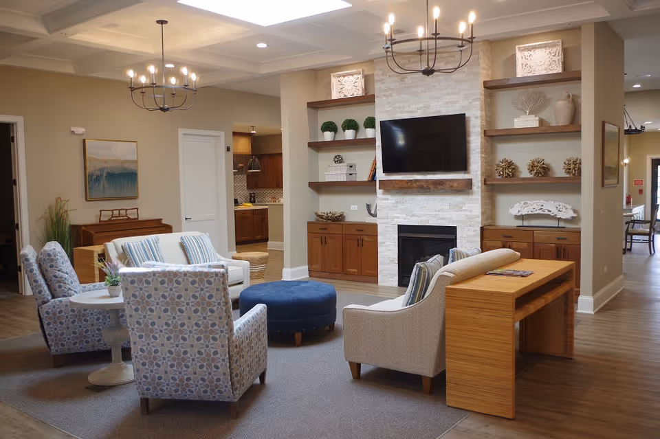 A cozy senior living room with a fireplace and a mounted flat-screen TV. The room features comfortable armchairs with patterned upholstery, a round blue ottoman, a wooden desk with a magazine on it, and built-in wooden shelves decorated with plants, books, and ornaments. The space is well-lit with ceiling lights and chandeliers, and the flooring is a combination of carpet and wood.
