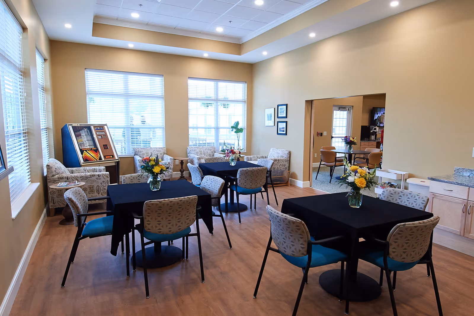 Bright communal dining room with several tables draped in black cloth, chairs, lounge seating by large windows, and floral centerpieces.