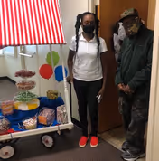 Two people wearing face masks stand indoors next to a small cart decorated with a red and white striped canopy. The cart holds various snacks and treats in clear containers and bags. The setting appears to be inside a facility hallway or room.