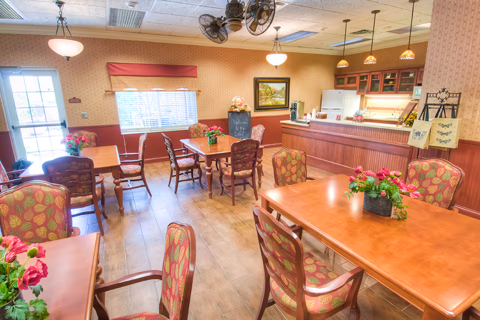 A cozy dining room in a retirement village with wooden tables and chairs featuring red and green leaf-patterned upholstery. Each table has a floral centerpiece. The room has warm beige wallpaper, a window with blinds and a red valance, and a counter area with a refrigerator and cabinets in the background. A chalkboard sign near the counter reads 'Welcome to the Café'.