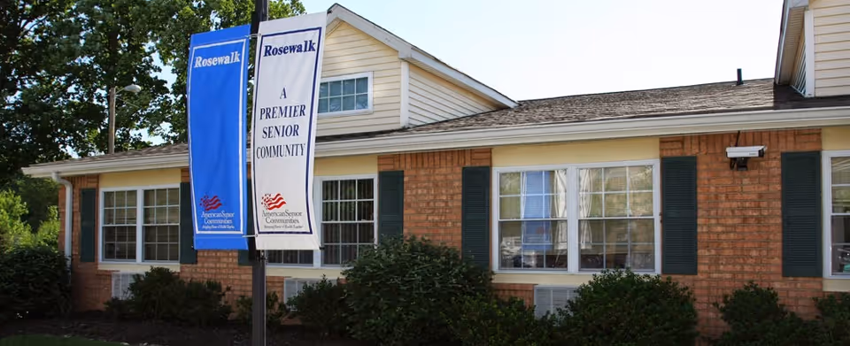 Exterior view of a single-story brick building with green shutters and multiple windows. Two vertical banners are displayed on a pole in front of the building, one blue and one white, both reading 'Rosewalk' and 'A Premier Senior Community' with the American Senior Communities logo. There are bushes and trees around the building.