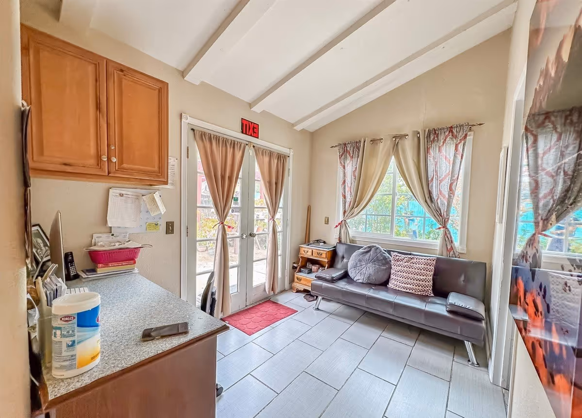A small, cozy room with a gray leather couch adorned with two decorative pillows, a wooden side table with a phone and other items, and a desk with cleaning wipes, a phone, and papers. The room has light-colored tiled flooring, beige walls, and a high ceiling with exposed white beams. There are two windows with patterned curtains and a set of glass double doors with beige curtains leading outside. A red exit sign is above the doors.