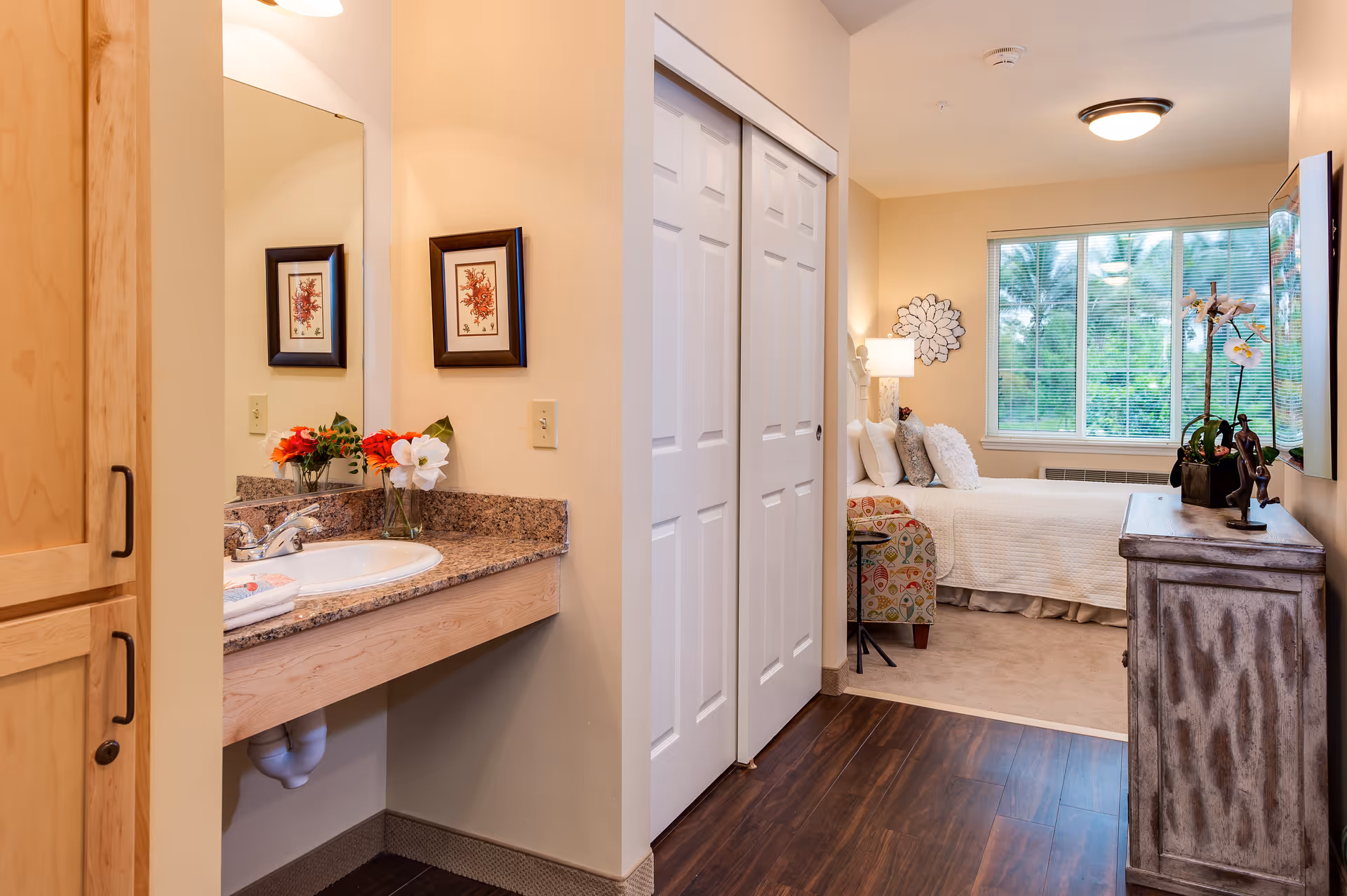 Interior view of a senior living facility room showing a small vanity area with a sink and mirror on the left, a hallway with white double closet doors, and a bedroom area with a bed, decorative pillows, a lamp, a window with blinds, and a wooden dresser with plants and decorative items on top.