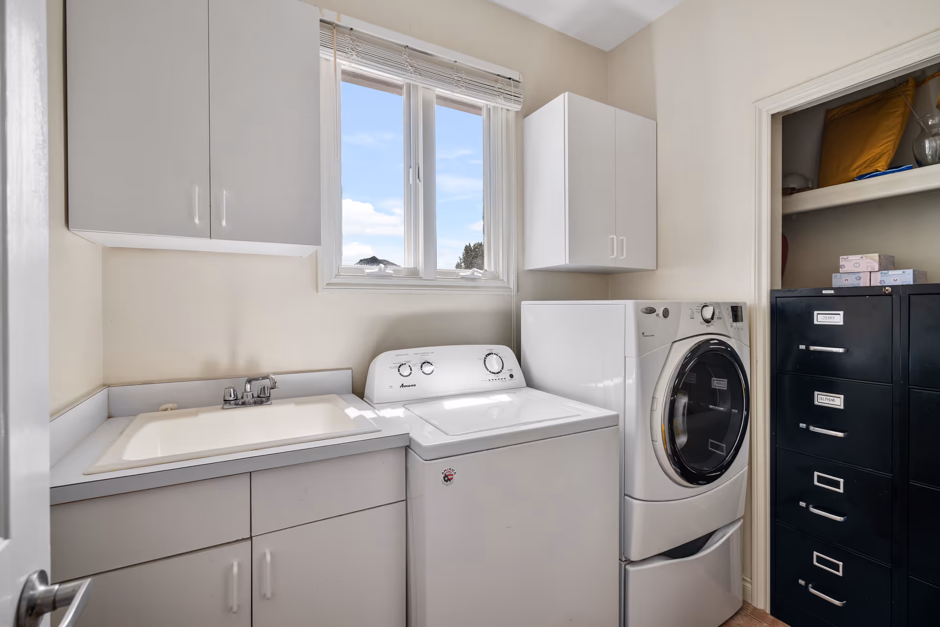 Laundry room with a white washing machine and dryer side by side under a window with blinds. There are white cabinets mounted on the walls above the machines and a sink with a faucet next to the washing machine. A black filing cabinet with labeled drawers is visible in an open closet to the right.