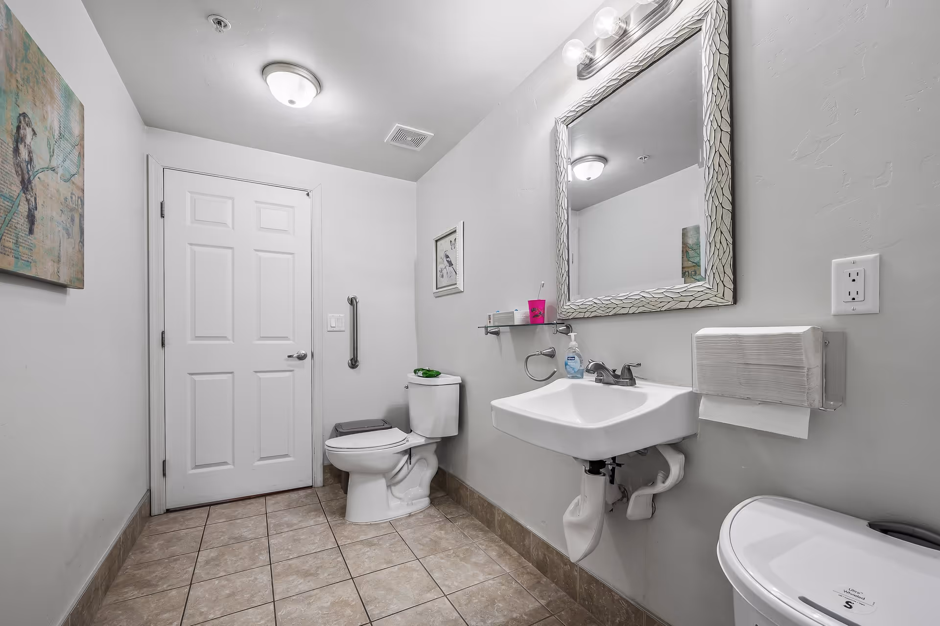 A clean, well-lit bathroom with a white door, a toilet with a grab bar beside it, a white sink with a mirror above, a soap dispenser, a pink cup, a paper towel dispenser, and two framed pictures on the walls. The floor is tiled and the walls are painted light gray.