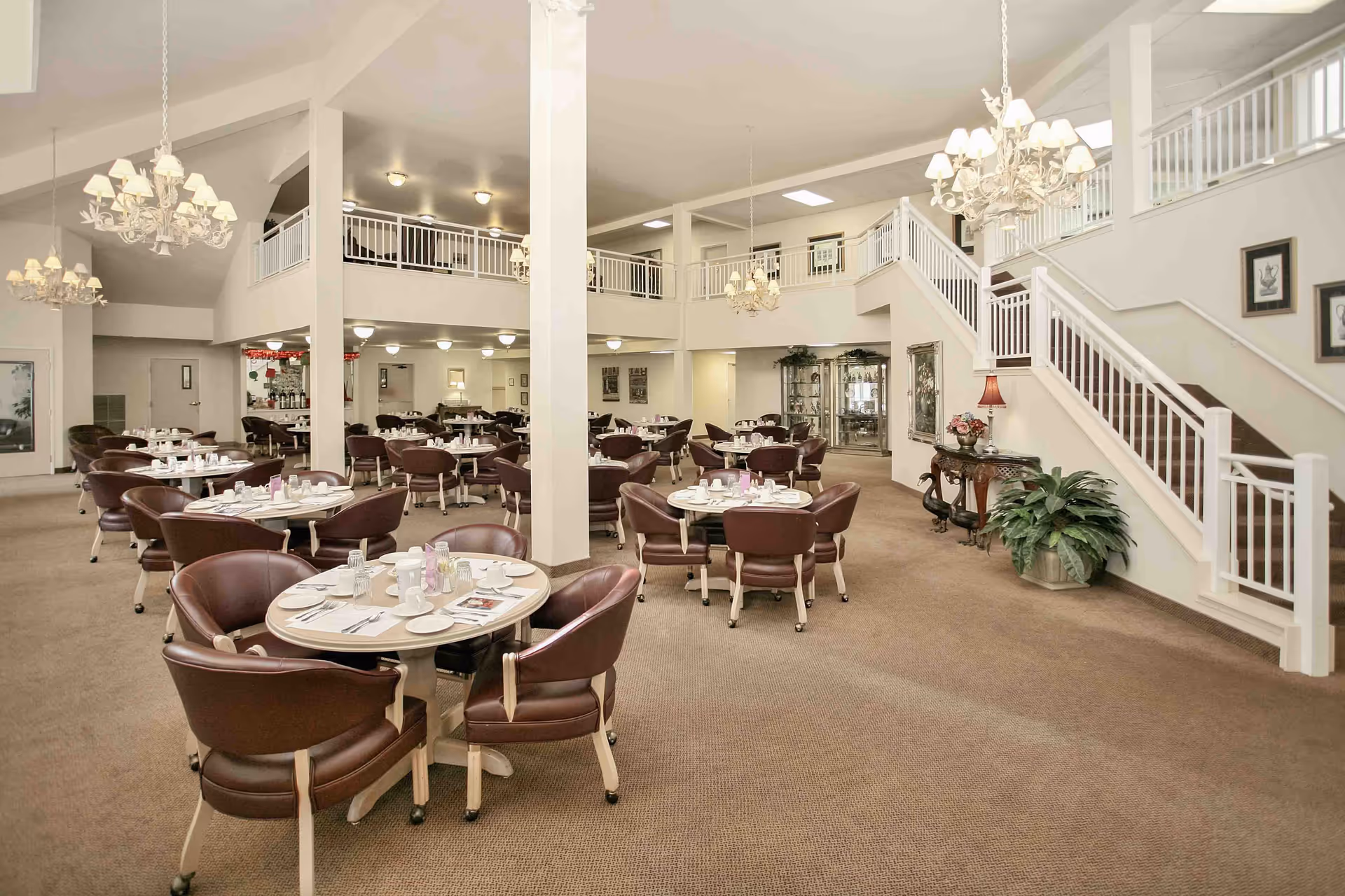 Spacious senior living dining room with multiple round tables set for meals, leather chairs, chandeliers, and a staircase leading to an upper balcony.