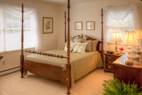 A cozy bedroom with a wooden four-poster bed covered with a beige quilt and multiple pillows. The room has two windows with white curtains, a wooden nightstand with a lamp and a clock, framed artwork on the walls, and a wooden dresser with two lamps and decorative items. A green plant is visible in the foreground.
