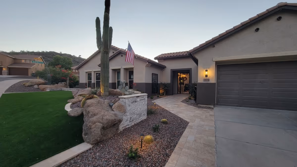 Exterior view of Anthem Senior Manor showing a single-story building with a garage, a walkway leading to the entrance, desert landscaping with rocks and cacti, and an American flag mounted near the front windows.