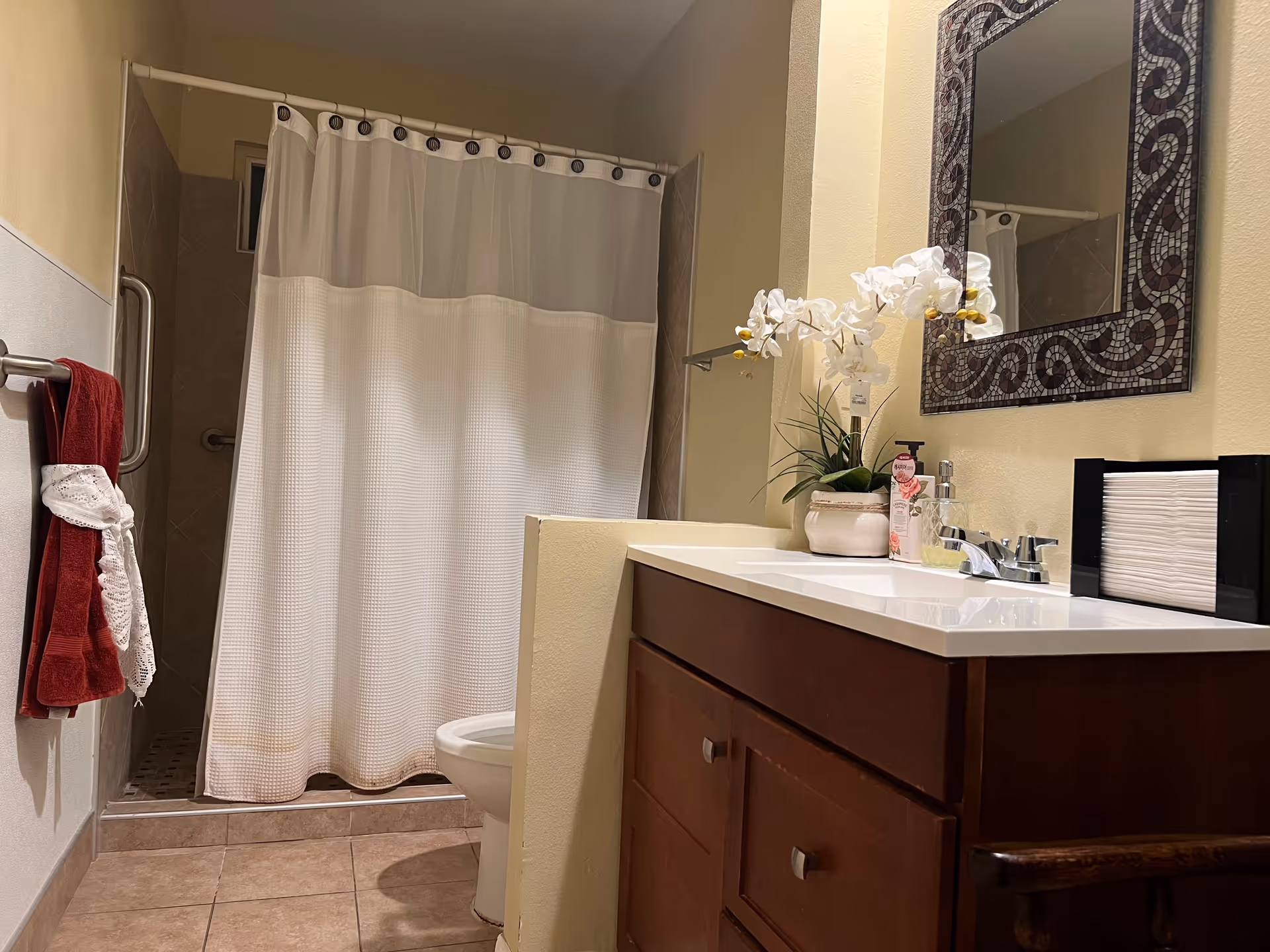 A bathroom with a tiled floor, a shower area with a white curtain, a toilet partially visible behind a half wall, a wooden vanity with a white countertop, a decorative mirror above the sink, a potted orchid plant, hand soap, and a black napkin holder. A red towel with a white decorative cloth is hanging on a towel rack on the left wall.