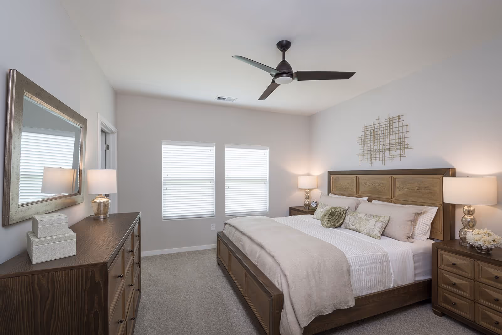 A neatly arranged bedroom with a large wooden bed featuring beige bedding and decorative pillows. There are two matching wooden nightstands on either side of the bed, each with a lamp. A wooden dresser with a large mirror above it is positioned against the opposite wall. Two windows with white blinds allow natural light into the room. A modern ceiling fan with three blades is mounted on the ceiling.