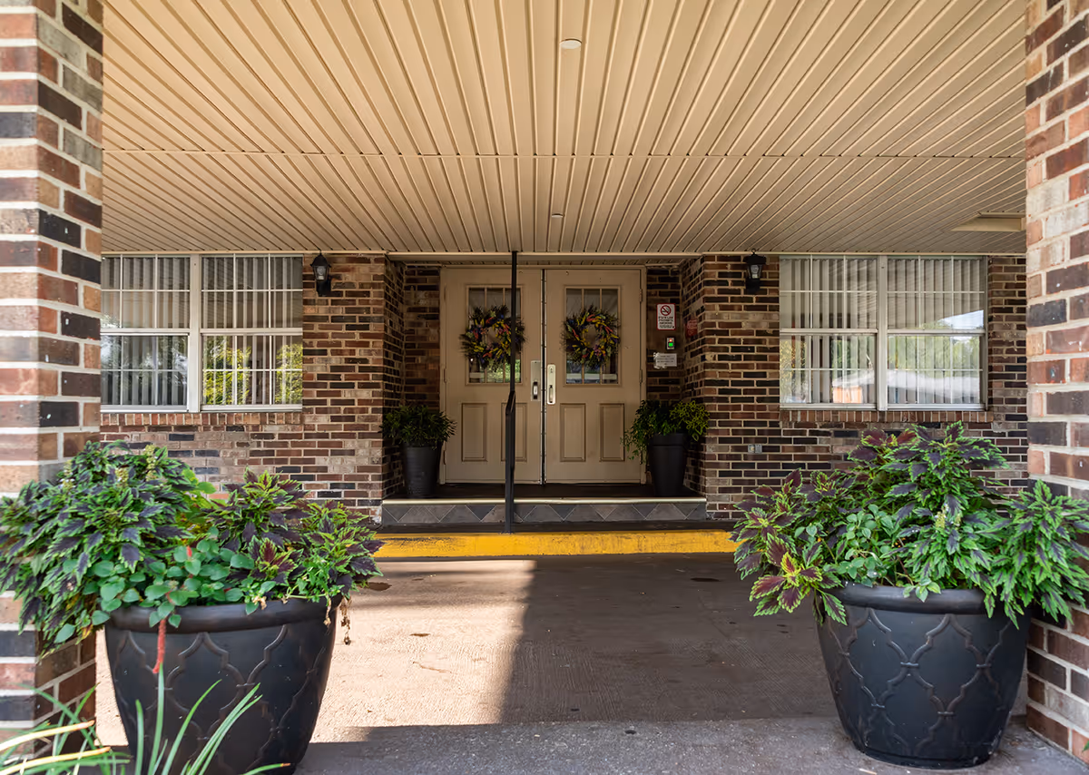 Covered entrance to a brick building with double doors decorated with wreaths, flanked by two large potted plants with green and purple foliage, and windows with white curtains on either side.