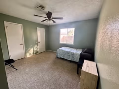 Sparsely furnished bedroom with a single bed, ceiling fan, window, and two doors.