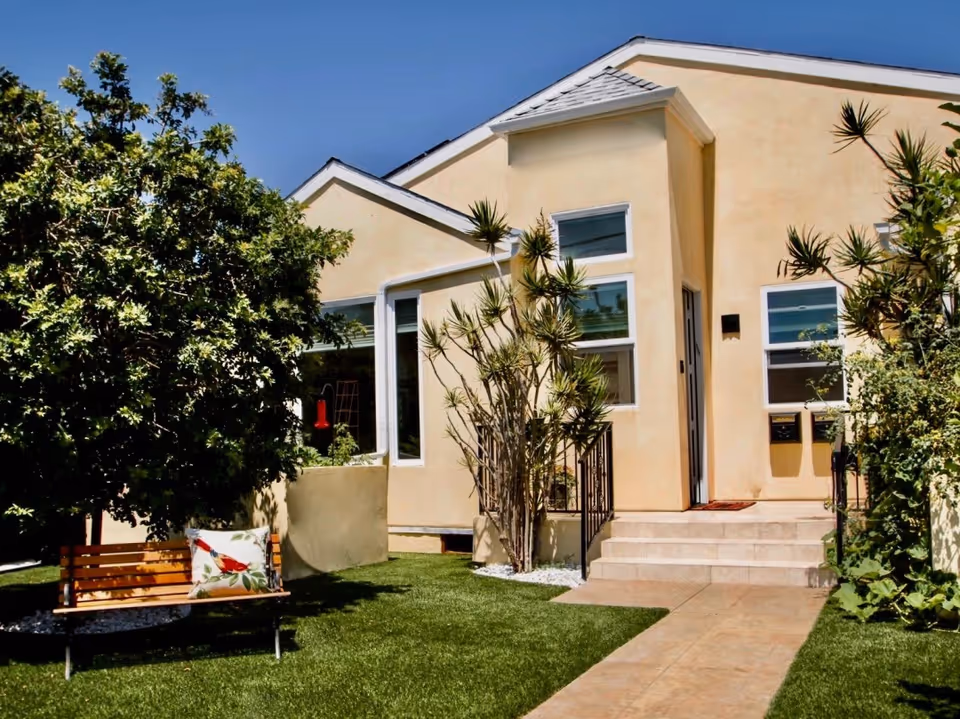 Exterior view of a beige building with a small staircase leading to a door, surrounded by green plants and trees. There is a wooden bench with a decorative pillow featuring a red bird on the left side on a well-maintained lawn under a clear blue sky.