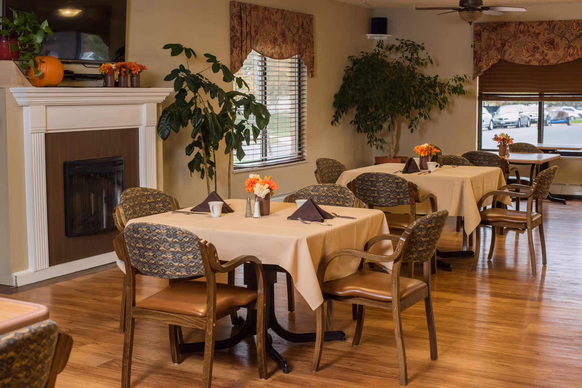 A cozy dining room with several tables covered in beige tablecloths, each set with napkins, cups, and small flower arrangements. The room features wooden chairs with patterned cushions, a fireplace with a pumpkin and plants on the mantel, large windows with blinds and valances, and indoor plants near the walls. The floor is wooden, and a ceiling fan is visible.