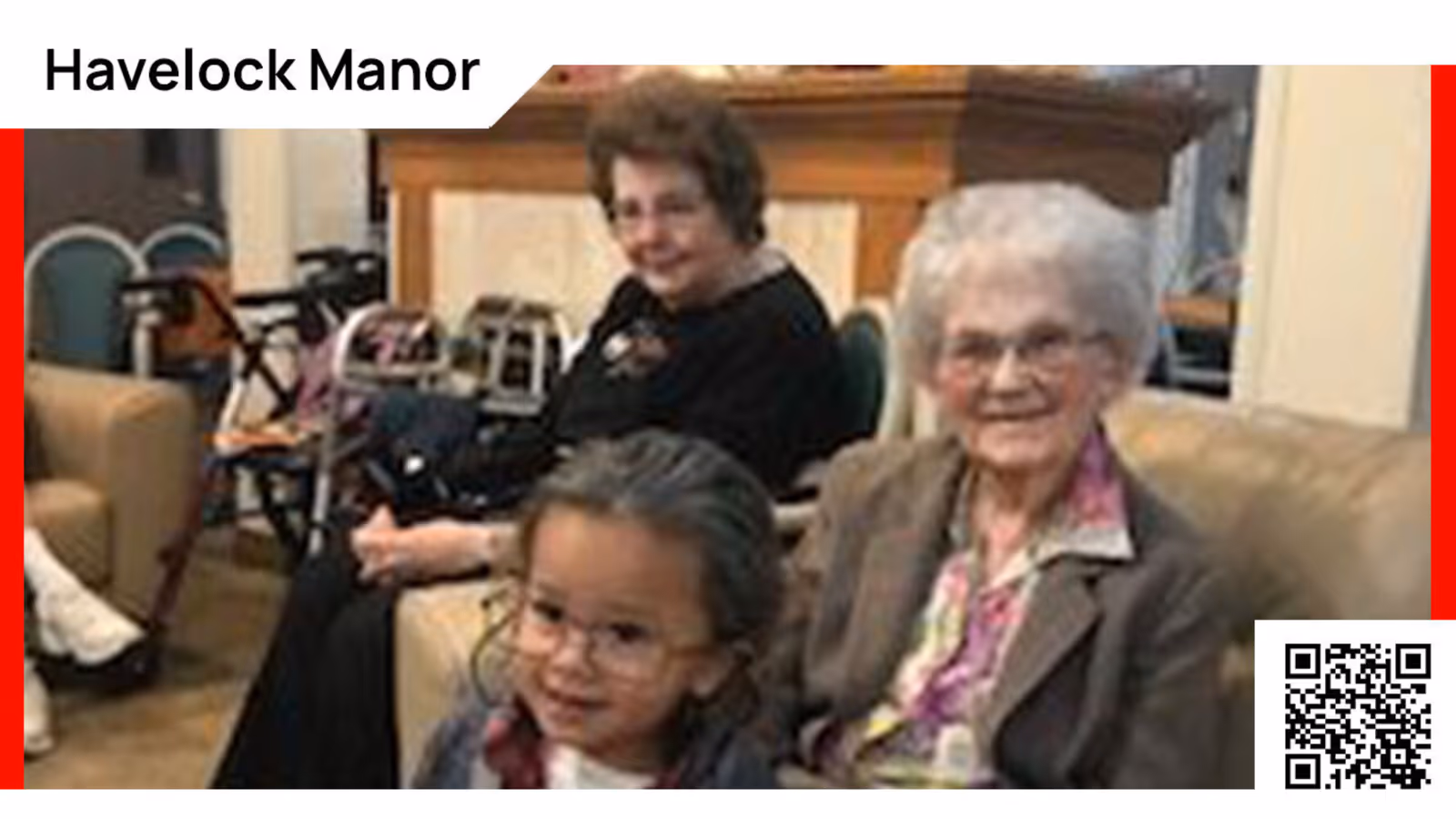 An elderly woman and a young girl sitting together on a couch in a living room setting, with another elderly woman sitting in the background. The room has chairs and mobility aids visible.
