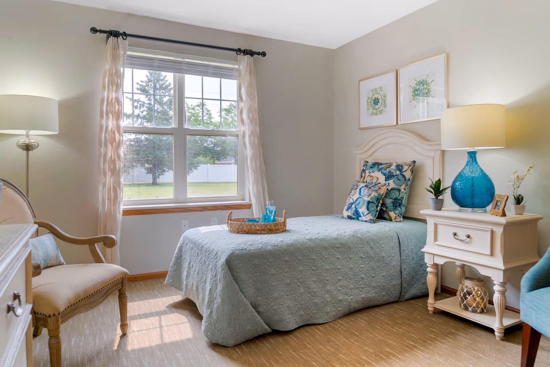 A bright and cozy bedroom with a single bed covered in a light blue quilt and floral pillows. A wicker tray with a glass and bowl sits on the bed. There is a wooden nightstand with a blue lamp, small plants, and a framed photo. A wooden chair with a cushion is placed near a large window with sheer curtains, which lets in natural light and shows a view of trees and a white fence outside. Two framed botanical prints hang on the wall above the bed.