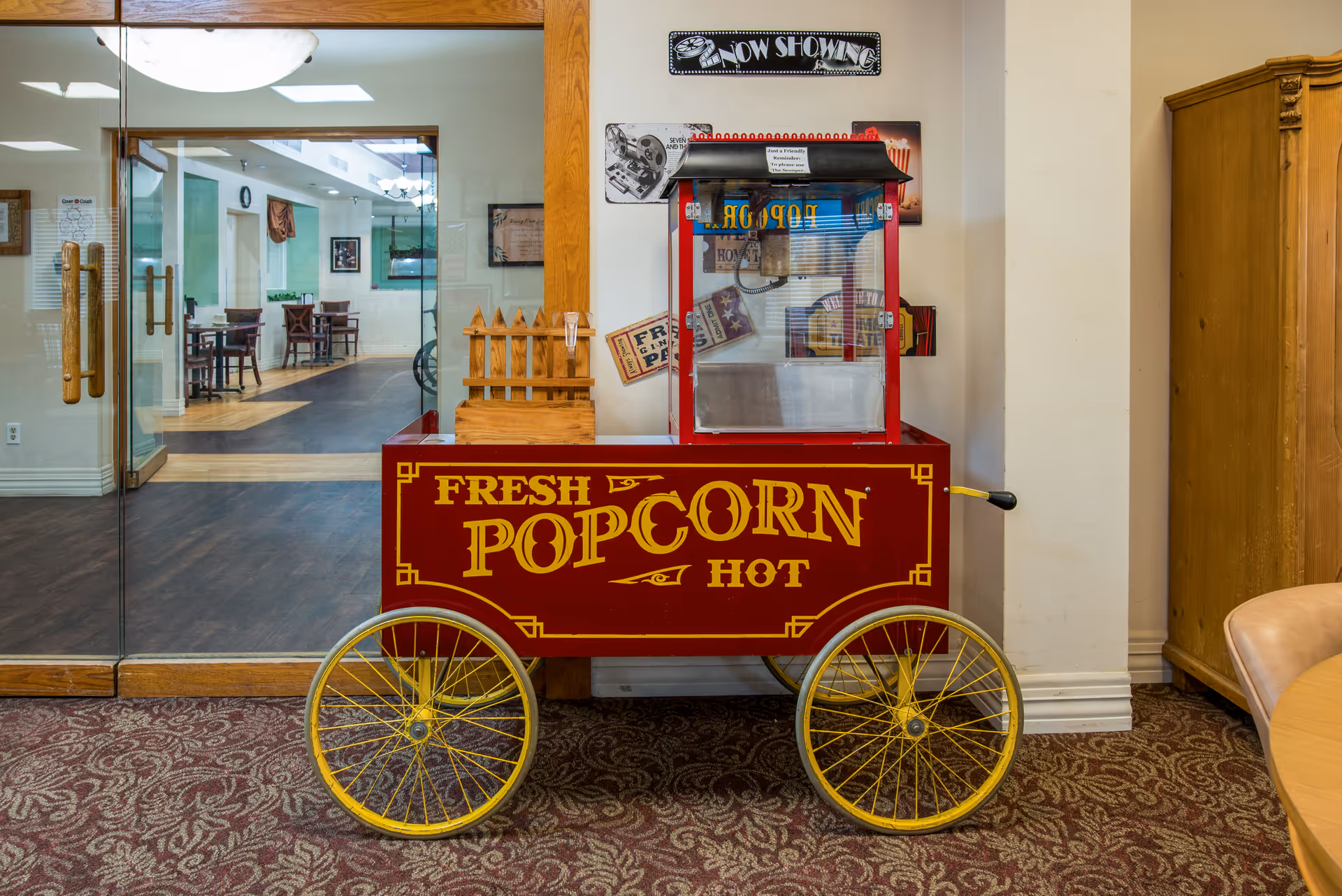 A vintage-style red and yellow popcorn cart with the words 'Fresh Popcorn Hot' written on it, positioned indoors on a patterned carpet. Behind the cart is a glass door leading to a dining area with tables and chairs. The wall behind the cart has movie-themed decorations including a 'Now Showing' sign.