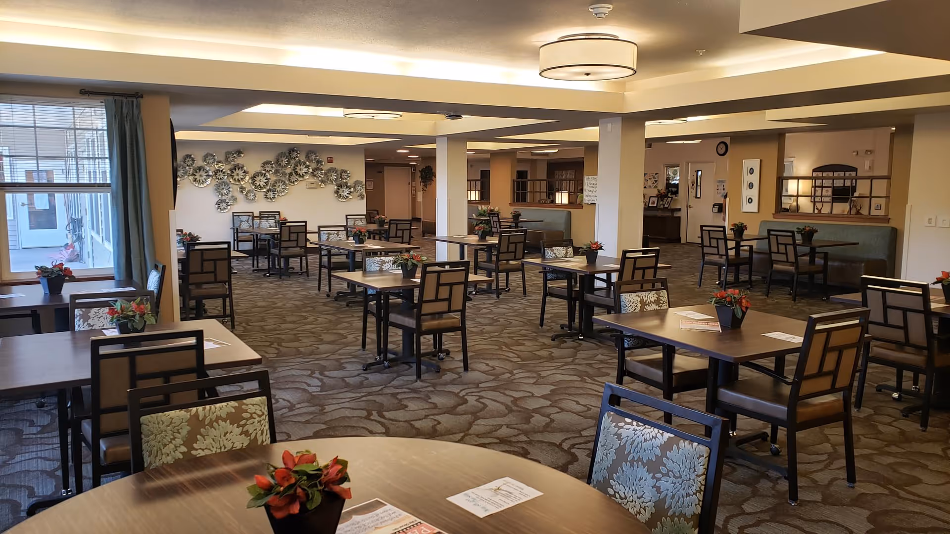 Spacious dining room in a senior living facility with multiple tables and chairs arranged neatly. Each table has a small potted plant with red flowers. The room has patterned carpet flooring, beige walls, and ceiling lights. Large windows with curtains allow natural light to enter the room. Decorative wall art is visible on the far wall.