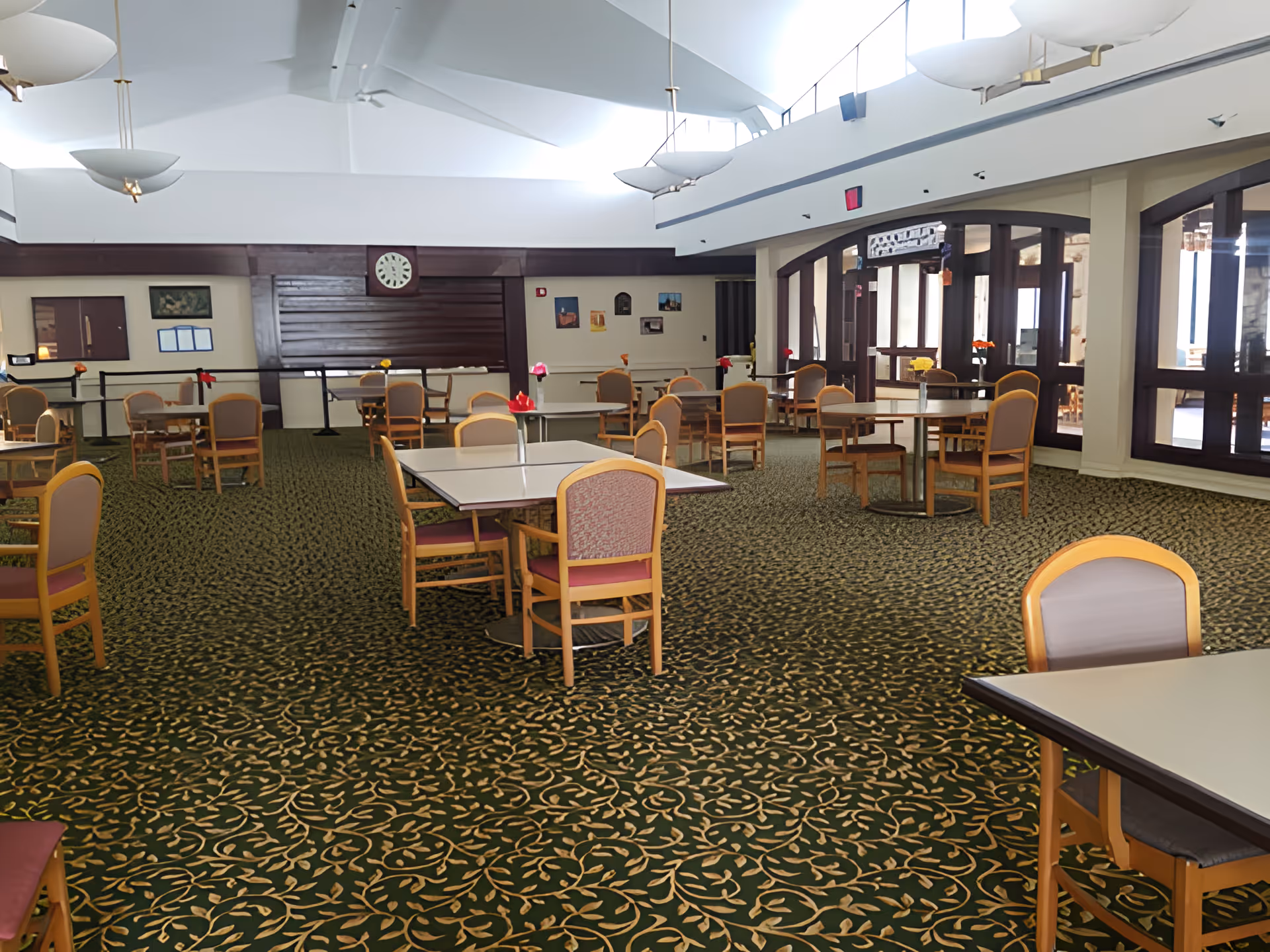 A spacious dining room with multiple tables and chairs arranged neatly on a patterned carpet. The room has large windows on one side allowing natural light to enter, and the ceiling features modern light fixtures. There are some framed pictures and a clock on the far wall.