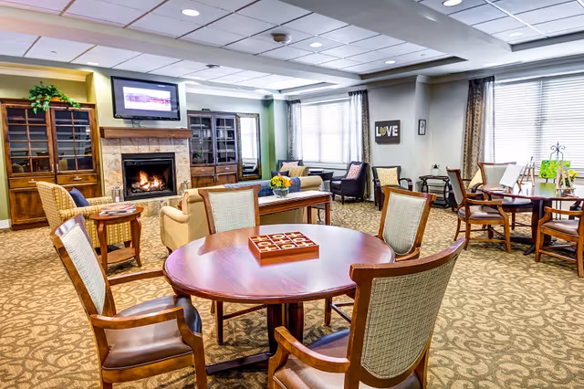 Bright, furnished senior living common room with round tables and chairs, lounge seating, a fireplace, and a wall-mounted TV.