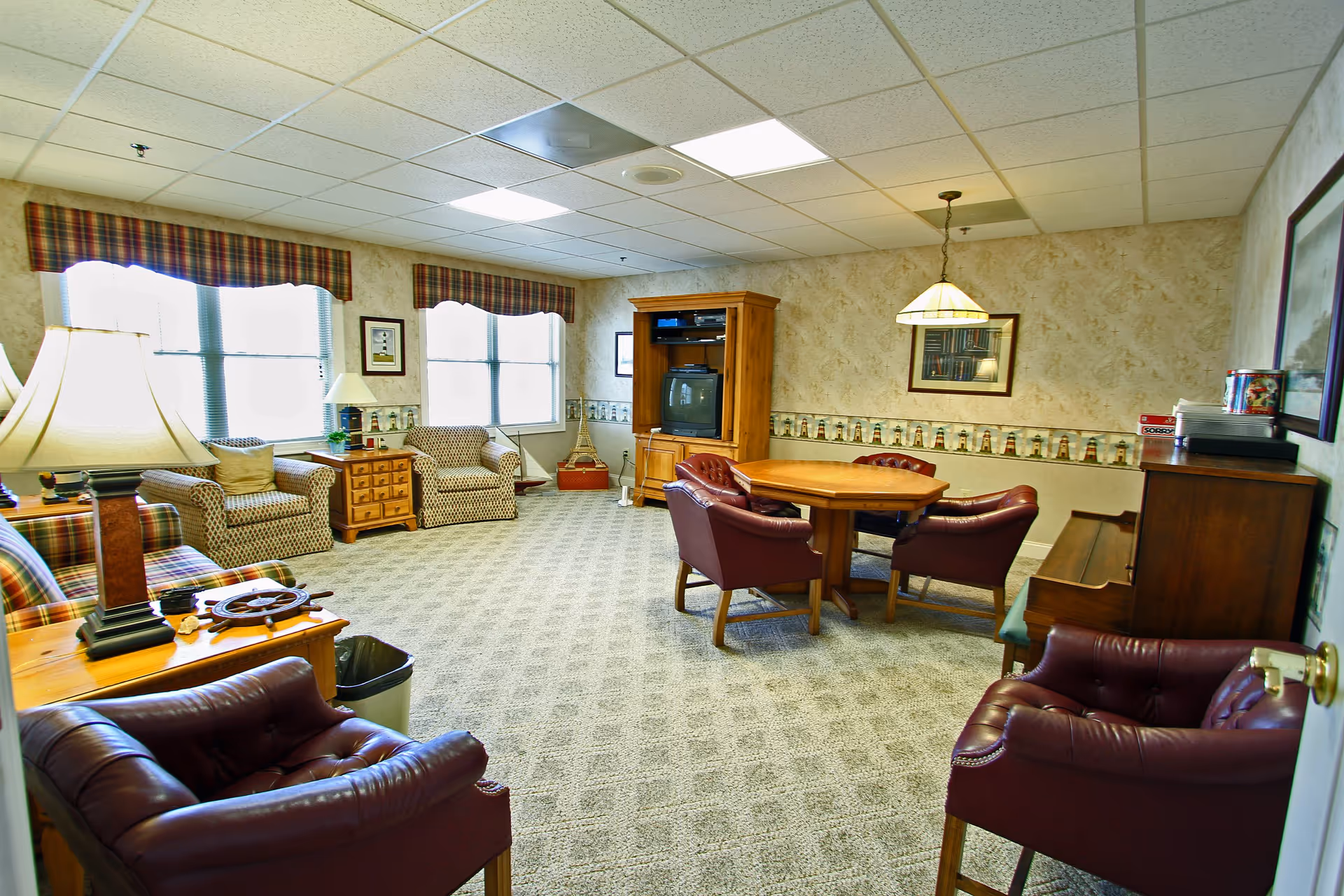 A cozy living room with patterned carpet and wallpaper, featuring a wooden entertainment center with a TV, a round wooden table surrounded by four leather chairs, two patterned armchairs near windows with plaid valances, a plaid sofa, a wooden side table with a lamp, and a piano against the wall.