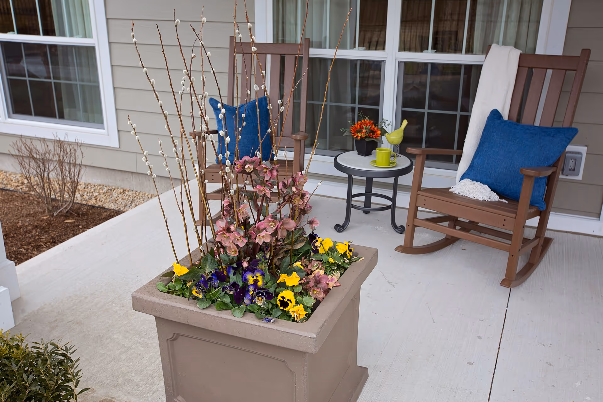 Outdoor patio area with two brown wooden rocking chairs, each with a blue cushion. One chair has a white throw blanket draped over it. Between the chairs is a small round table with a green cup, a small flower arrangement, and a decorative yellow bird. In the foreground is a large square planter filled with colorful flowers and tall branches. The background shows windows and beige siding of the building.