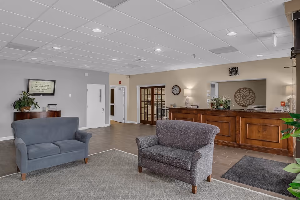 A spacious and well-lit reception area with two gray upholstered loveseats on a carpeted floor. Behind the loveseats is a wooden reception desk with plants and a lamp. On the wall behind the desk is a clock and a decorative wall piece. To the left, there is a small wooden cabinet with a plant and a framed item on top, and a wall-mounted screen displaying information. The area has a tiled floor and a drop ceiling with recessed lighting.
