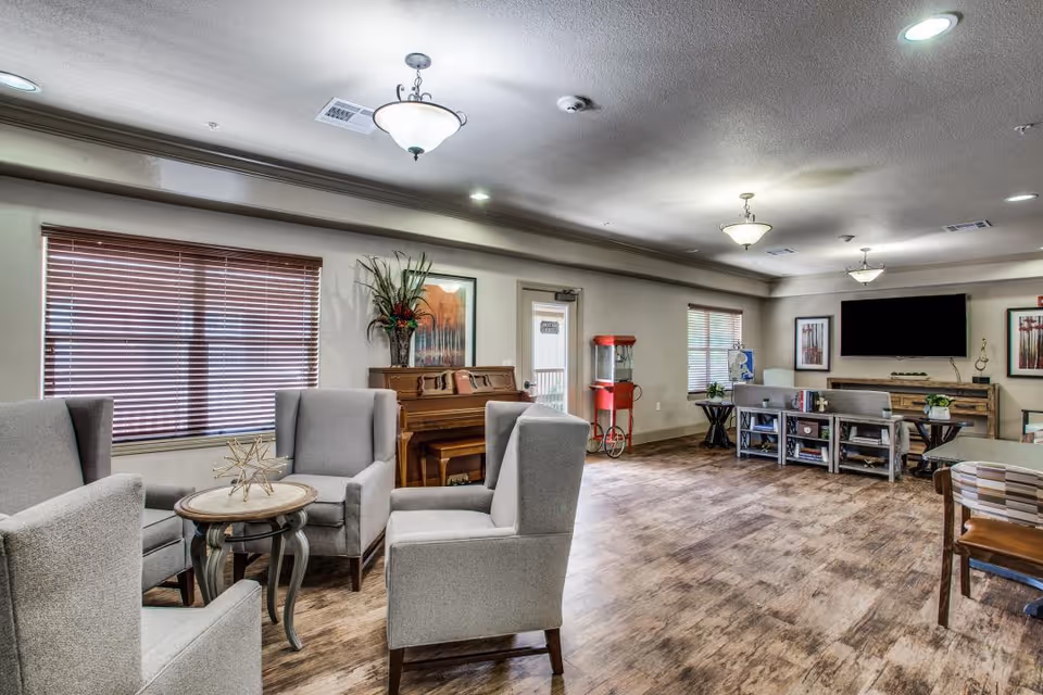 Spacious senior living community lounge with armchairs around a small table, a piano, popcorn machine, TV, and wood-look flooring.