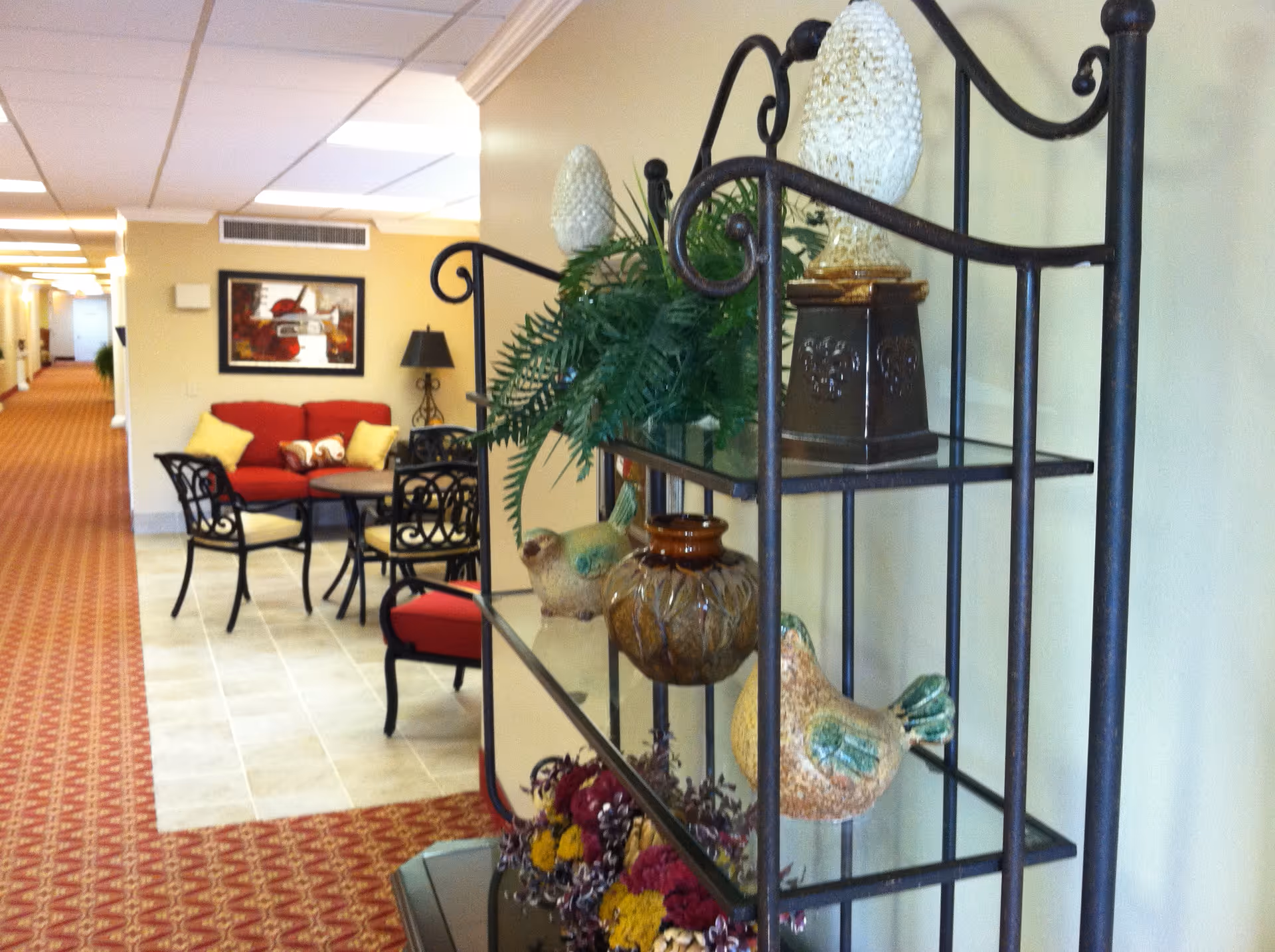 A hallway in a senior living facility with a decorative metal shelf holding ceramic birds, a vase, and plants in the foreground. In the background, there is a seating area with a red couch, black metal chairs with yellow cushions, a round table, a floor lamp, and a framed picture on the wall. The floor is carpeted with a red patterned carpet, and the walls are painted light yellow.