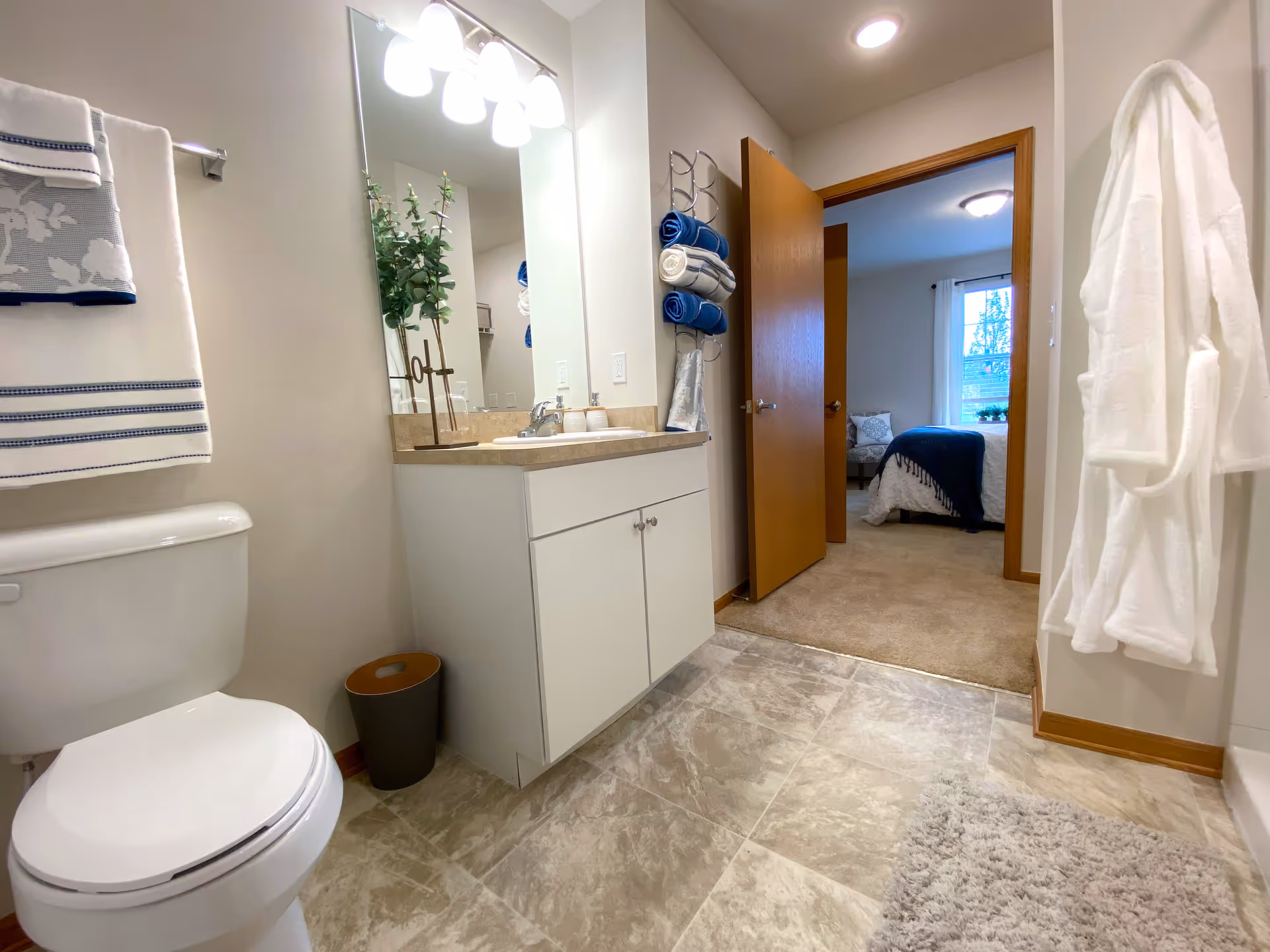 Bright bathroom with a vanity and mirror, toilet, towels and robe, and an open door leading to a bedroom.