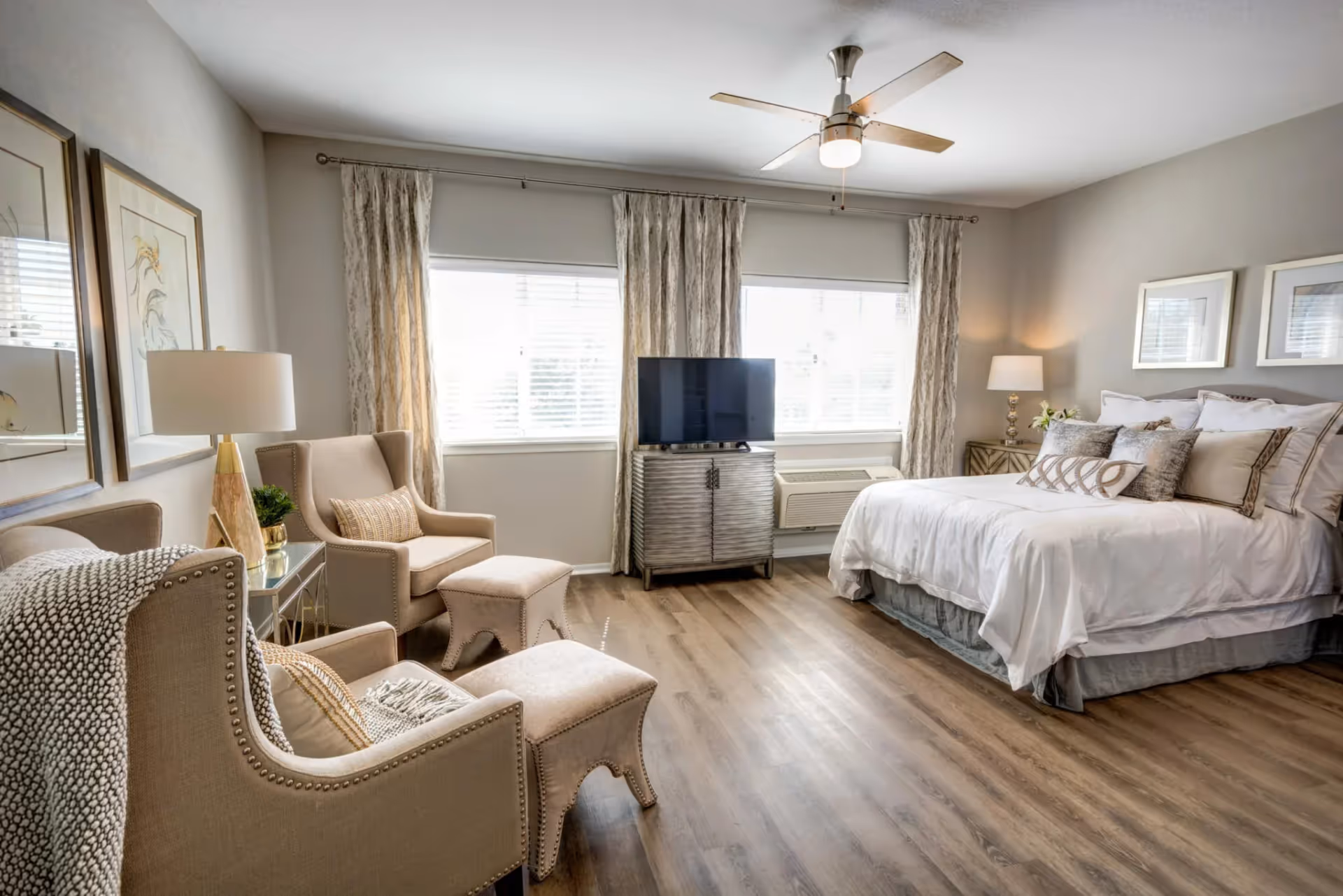 A bright and spacious bedroom with a large bed dressed in white linens and multiple pillows. Two beige armchairs with matching ottomans are positioned near a window with light-colored curtains. A flat-screen TV sits on a wooden cabinet between the windows. The room features wood flooring, a ceiling fan with light, and soft lighting from table lamps on either side of the bed.