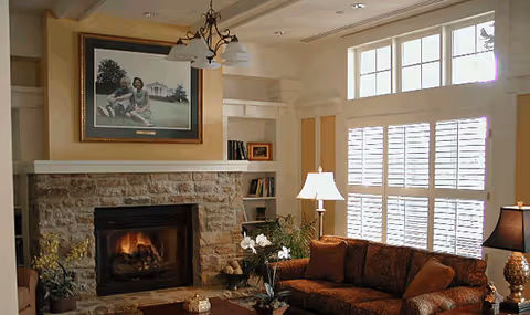 Cozy living room with a stone fireplace, a lit fire, a large framed family photo above the mantel, built-in shelves with books and decor, a brown patterned sofa with cushions, a coffee table with flowers, and large windows with white shutters letting in natural light.