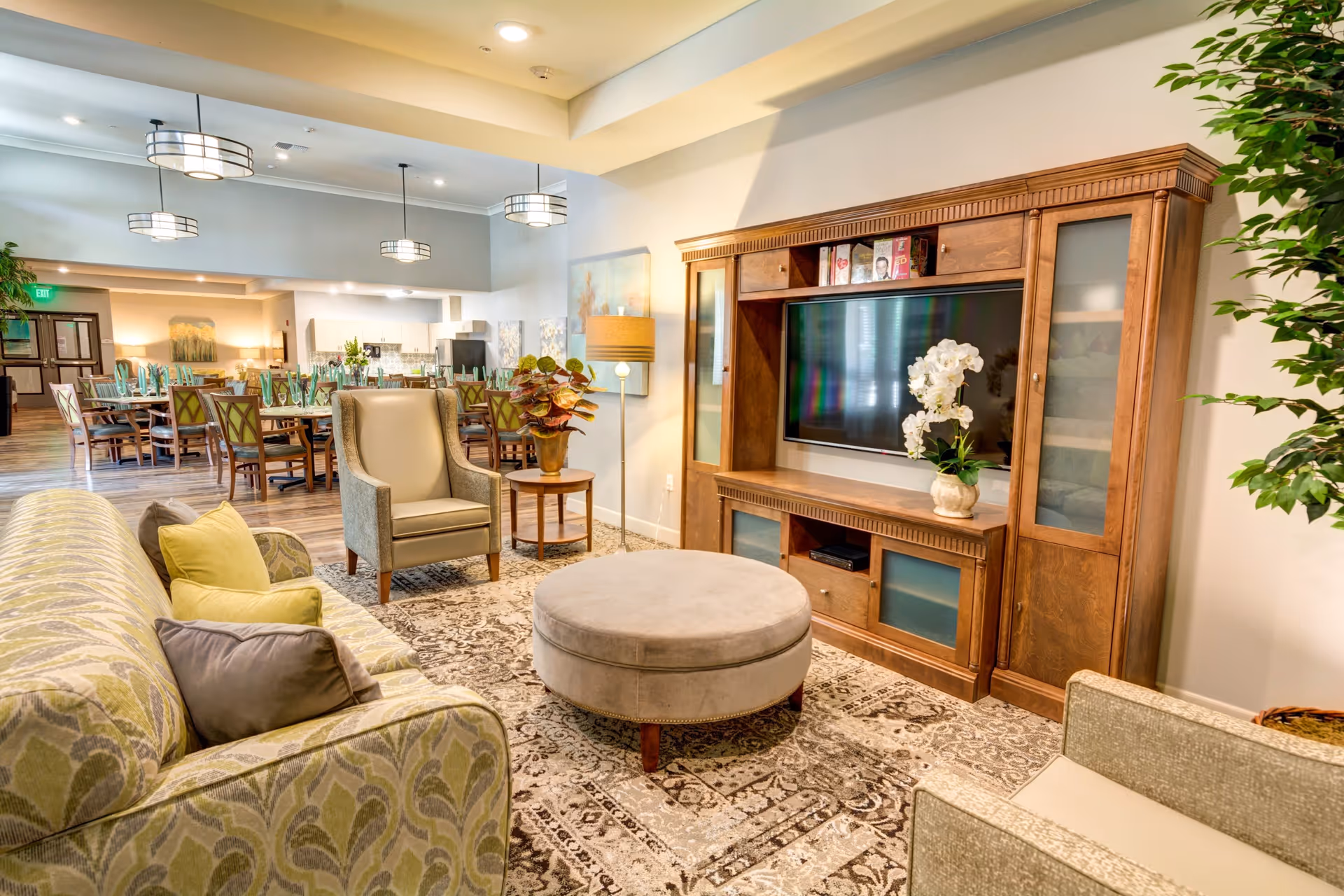 A cozy living room area with a patterned sofa, armchairs, a round ottoman, and a large wooden entertainment center with a flat-screen TV and decorative plants. In the background, there is a dining area with tables and chairs set for a meal, illuminated by modern pendant lights.