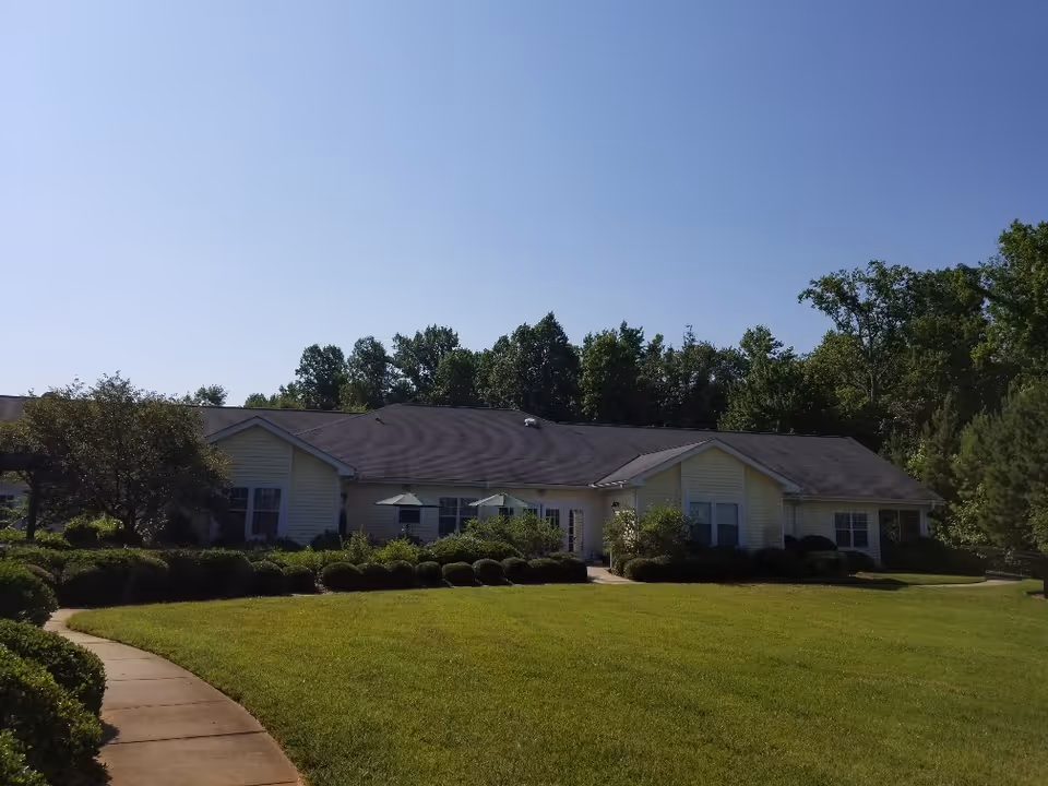 Single-story building with a dark roof and light-colored siding surrounded by green bushes and trees under a clear blue sky. A curved concrete walkway leads to the entrance.