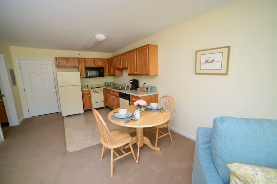 A small kitchen and dining area in a senior living facility apartment. The kitchen has wooden cabinets, a refrigerator, stove, microwave, and dishwasher. In front of the kitchen is a round wooden table set for two with plates, bowls, cups, and a small flower arrangement. A blue couch is partially visible on the right side, and a framed picture hangs on the wall above the table.