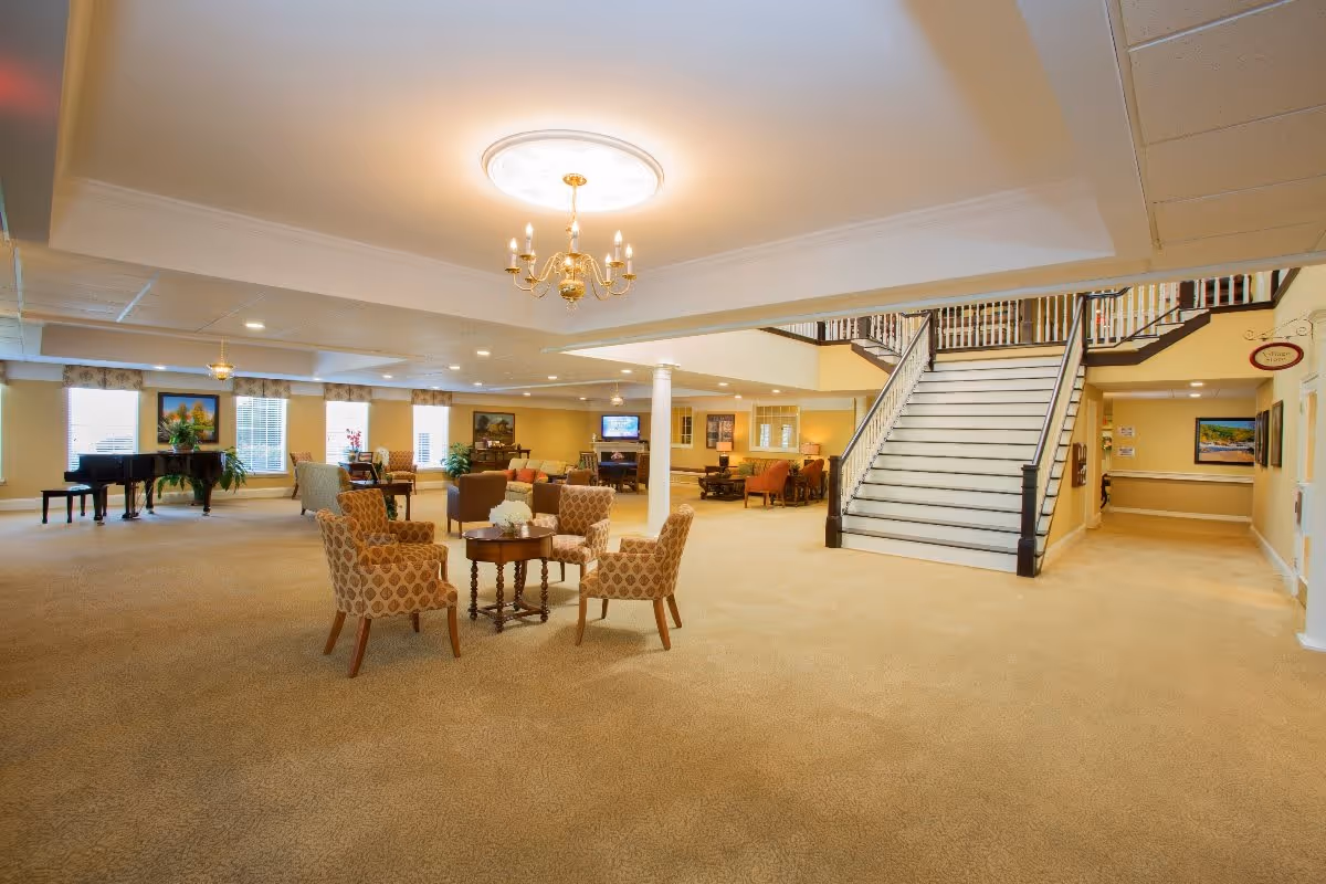 Spacious and well-lit common area in a senior living facility featuring a central seating arrangement with four patterned armchairs around a small wooden table. The room has beige carpeting, a grand staircase leading to an upper floor, a chandelier hanging from the ceiling, and additional seating areas with sofas and chairs. Large windows allow natural light to fill the space, and a grand piano is visible in the background.
