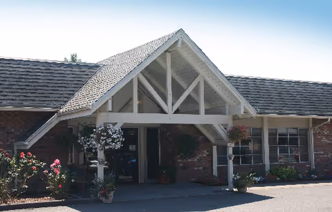 Front entrance of a brick senior living facility with a covered gabled portico, potted plants and flowering shrubs.
