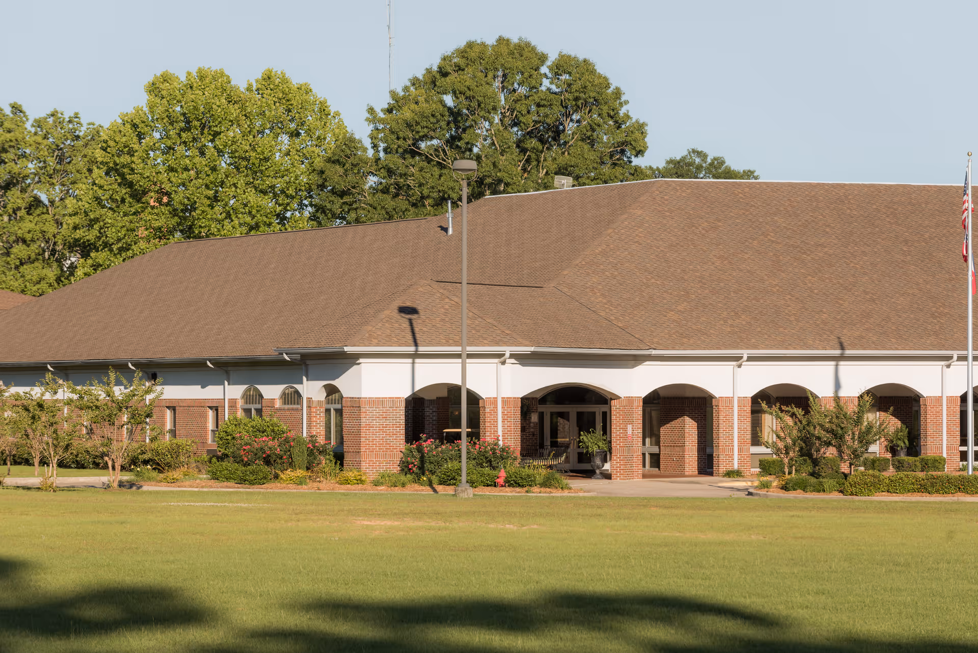 Front exterior of a single-story brick senior living facility with arched entrances, landscaping, and a flagpole on a grassy lawn.