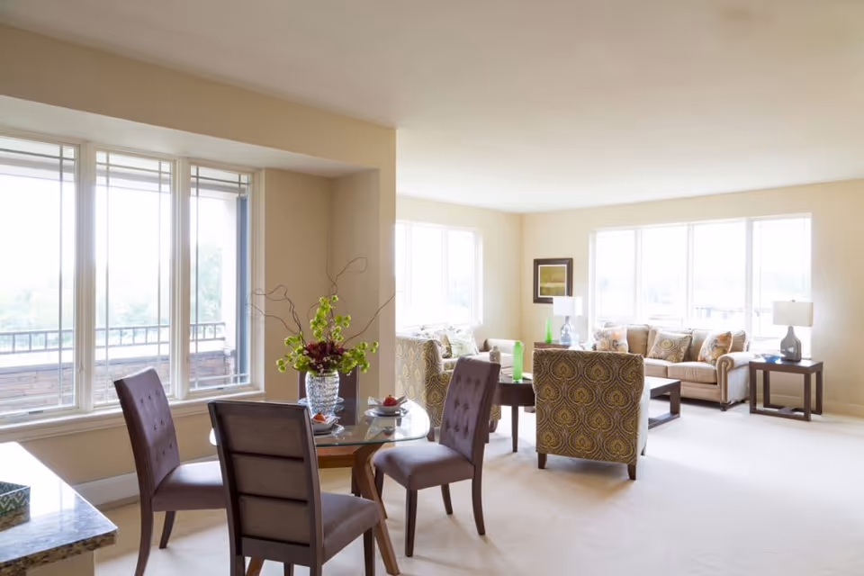 Bright open living and dining area with large windows, sofas, patterned chairs, and a glass dining table with a floral arrangement.
