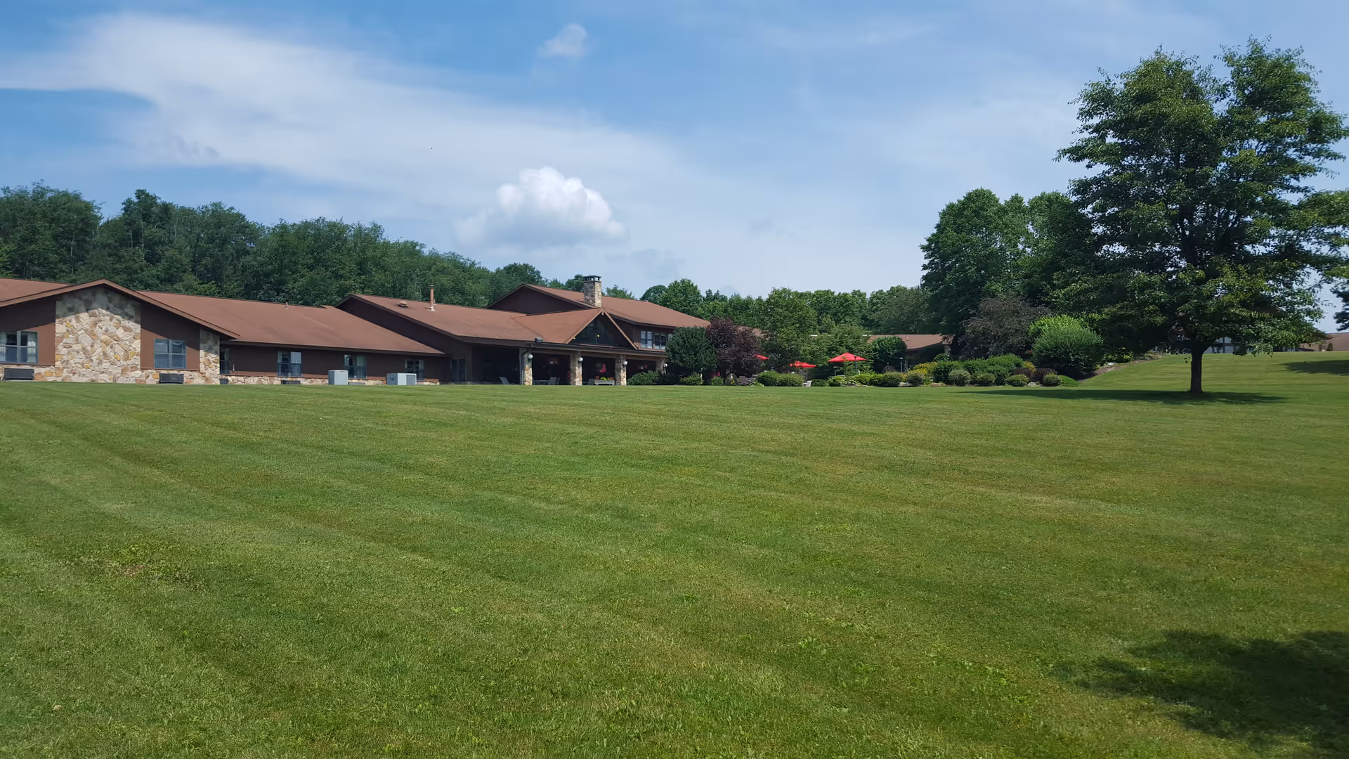 A low, single-story brown-roofed building sits beyond a wide mowed lawn with trees and red patio umbrellas.