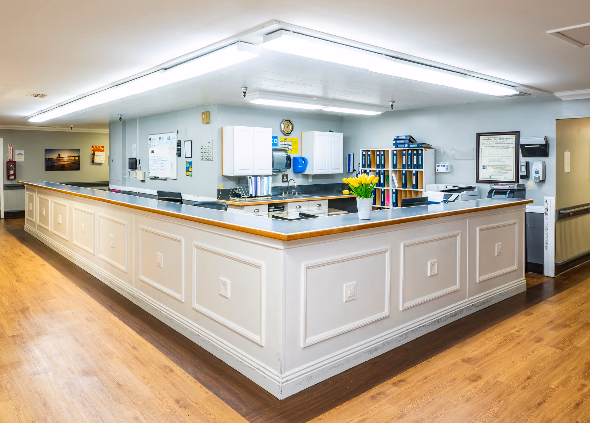 A clean and well-lit nursing center reception desk area with white paneled counters, wooden flooring, and overhead fluorescent lighting. The desk has office supplies, a vase with yellow tulips, file organizers, and cabinets on the wall behind it. There are informational posters and a clock on the walls.