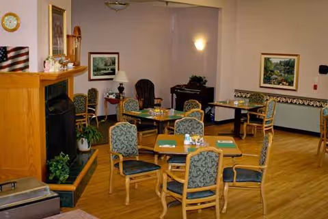A senior living facility dining area with green upholstered chairs around wooden tables and fireplace.