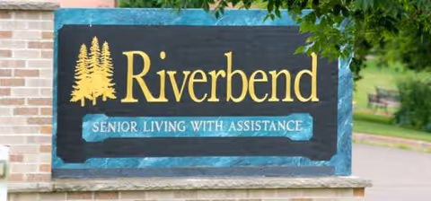Sign reads Riverbend Senior Living with Assistance mounted on a brick wall outdoors, with trees nearby.