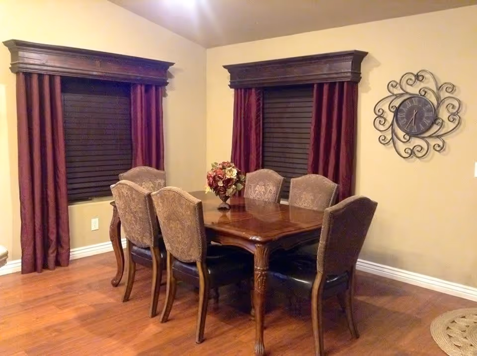 A dining room with a wooden dining table surrounded by six upholstered chairs. The room has two windows with dark blinds and burgundy curtains with wooden valances. A decorative wall clock with an ornate metal frame hangs on the beige wall. A floral centerpiece is placed on the table.
