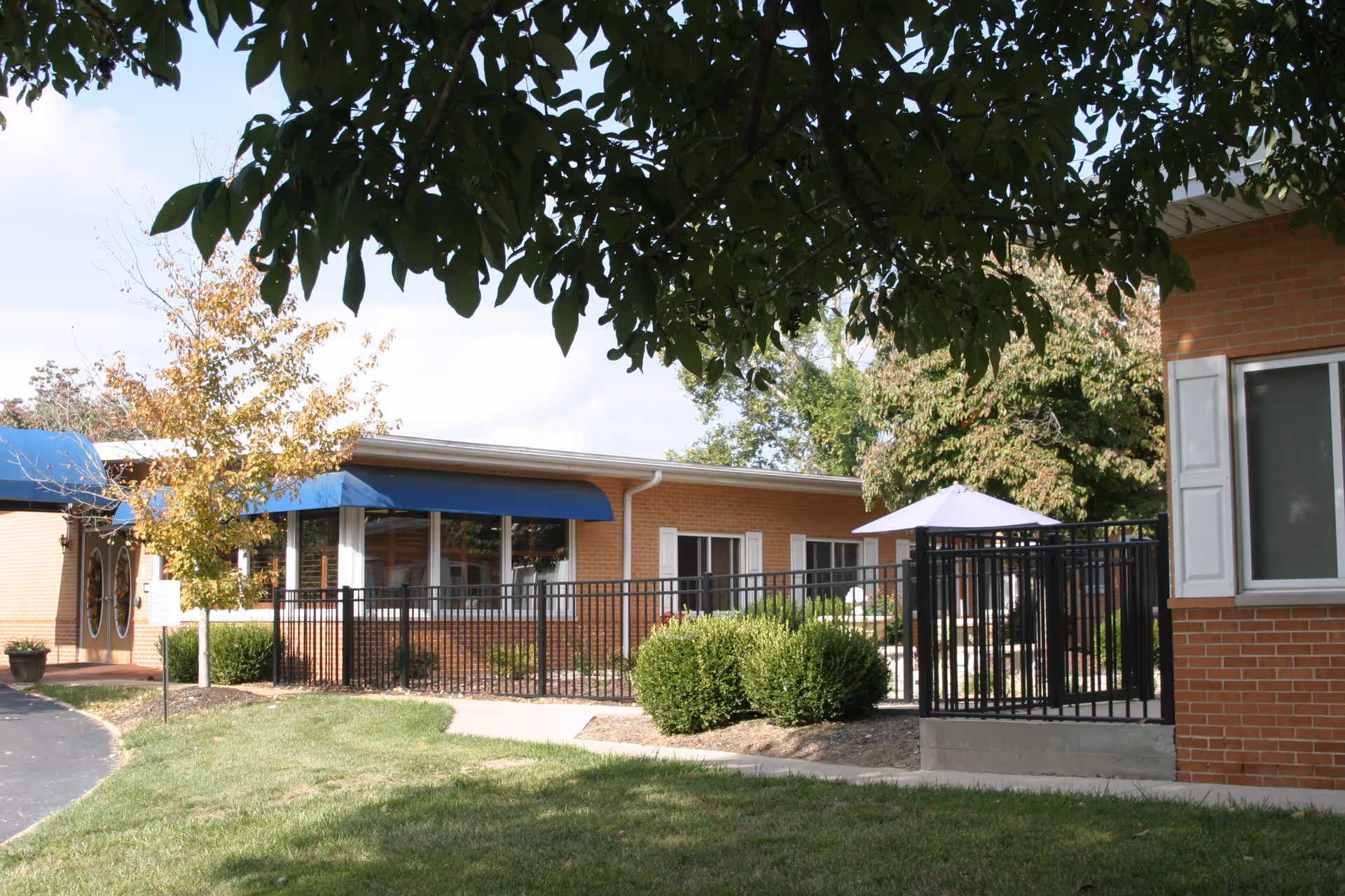Outdoor view of Sycamore Village Assisted Living showing a brick building with blue awnings over windows and doors, a black metal fence enclosing a patio area with a purple umbrella, surrounded by green grass, bushes, and trees.