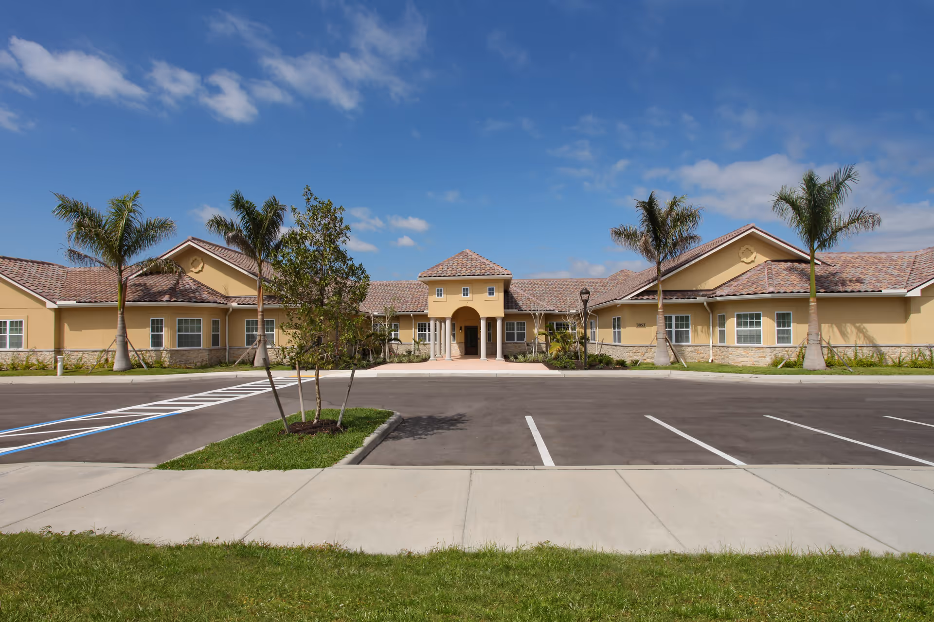 Front exterior view of The Fairways at Naples facility with a beige building featuring a tiled roof, palm trees, a small landscaped area, and an empty parking lot under a partly cloudy blue sky.